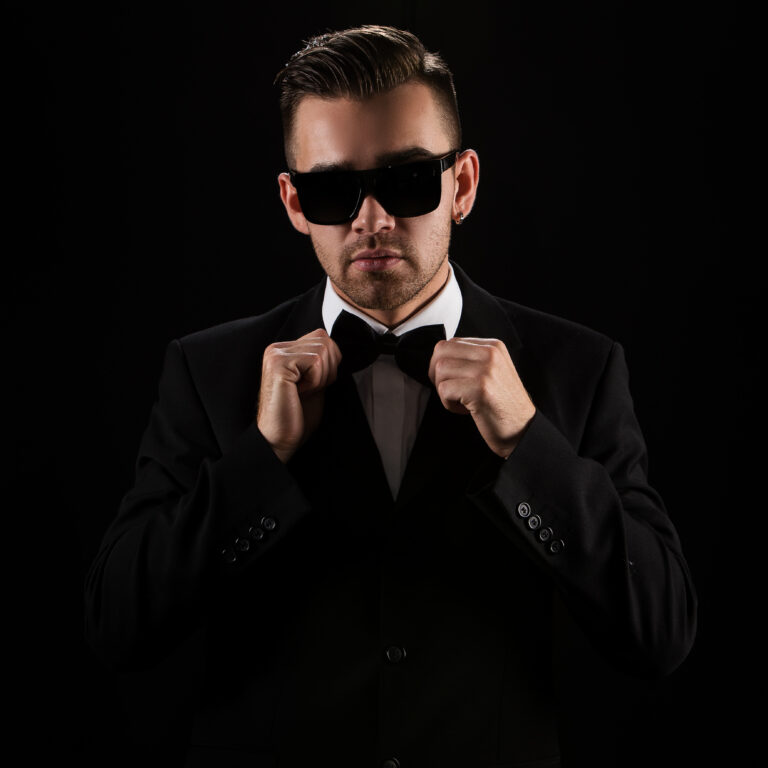 La imagen presenta un hombre joven vestido en un esmoquin de color negro con gafas negras y la corbata de lazo de color negro en un ambiente oscuro.