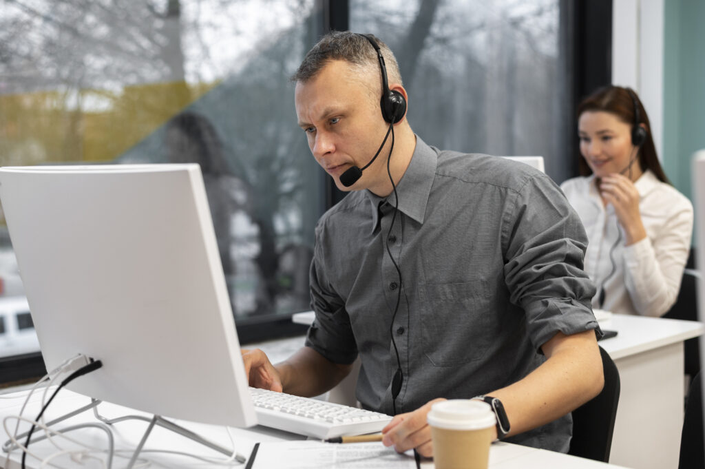 La imagen presenta a un hombre trabajando en el call-center. El hombre lleva una camisa de color gris oscuro, unos cascos con micrófono, se encuentra en una oficina de tarde. Utiliza un conjunto de ordenador y periféricos de color blanco y tiene a su izquierda un café.